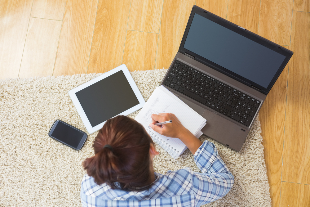 Brunette young student using her tablet and notebook for doing homework lying in the living room