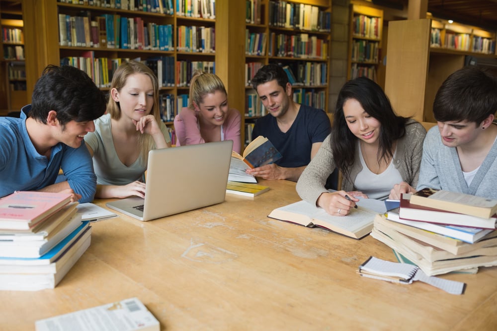 Students sitting at a table in a library while learning and working on a laptop-1