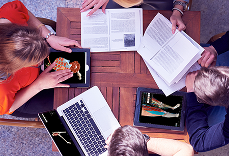 4 students studying around a table with textbooks open, 2 tablets and a laptop all with Visible Body 3d anatomy on the screens