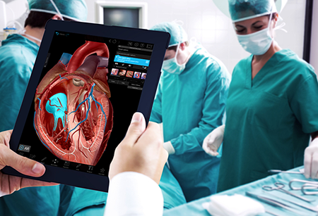 Visible Bodyの3D解剖学的構造が表示されているタブレットを持つ人物と、その後ろで作業する外科医たち